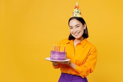 Happy Fun Smiling Young Woman Wearing Casual Clothes Cap Hat Celebrating Holding In Hand Purple Cake With Candles Look Camera Isolated On Plain Yellow Background. Birthday 8 14 Holiday Party Concept