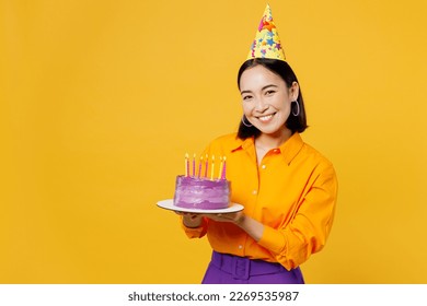 Happy fun smiling young woman wearing casual clothes cap hat celebrating holding in hand purple cake with candles look camera isolated on plain yellow background. Birthday 8 14 holiday party concept