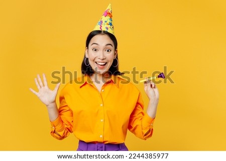 Happy fun surprised shocked young woman wearing casual clothes celebrating hold in hand blowing pipe look camera spread arms isolated on plain yellow background. Birthday 8 14 holiday party concept