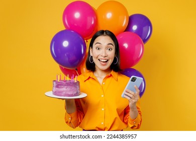 Happy fun surprised young woman wearing casual clothes celebrating near balloons hold cake with candles use mobile cell phone isolated on plain yellow background. Birthday 8 14 holiday party concept - Powered by Shutterstock
