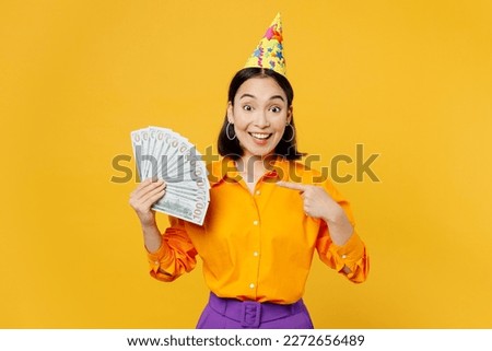 Happy fun surprised young woman wears casual clothes hat celebrating hold in hand point on fan of cash money in dollar banknotes isolated on plain yellow background Birthday 8 14 holiday party concept