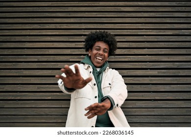 Happy funky gen z hipster rapper African American guy dancing, singing, having fun feeling hip-hop or rap vibe, moving and gesturing standing at wooden wall background outdoors.