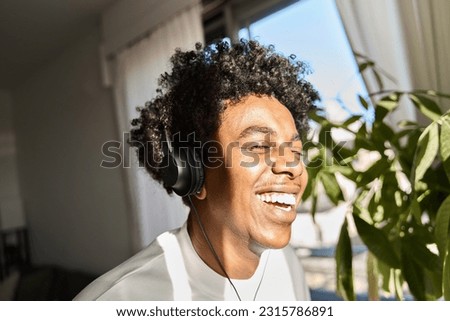 Happy funky gen z hipster African American guy wearing headphones laughing at home, listening music, having fun feeling relaxed standing in living room at home. Close up authentic shot.