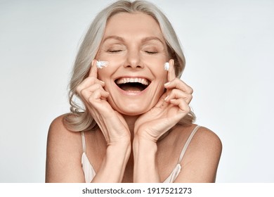 Happy Funny 50s Middle Aged Mature Old Blond Woman Applying Facial Cream On Face Laughing Enjoying Skin Care Beauty Spa Treatment Creme Isolated On White Background. Mid Age Skincare Concept.