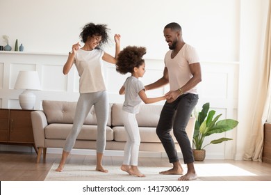 Happy funny active african family with cute little kid daughter dancing at home, carefree cheerful black parents mom dad and small child girl having fun jumping laughing enjoy leisure in the morning