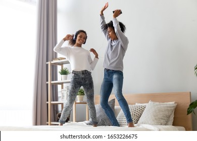 Happy funny african american family mom sister and teen daughter listening to music in headphones dancing on bed, excited active black mother with teenage girl jumping playing having fun in bedroom
