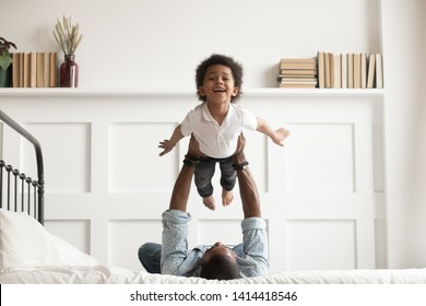 Happy funny african american kid boy flying in fathers arms looking at camera in bedroom, loving family single black dad holding lifting cute little child son playing plane bonding having fun on bed