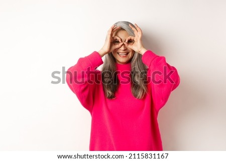 Happy and funny asian senior woman making hand binoculars, looking through fingers and smiling, standing in pink sweater over white background