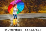 Happy funny ba child by girl with a multicolored umbrella jumping on puddles in rubber boots and laughing
