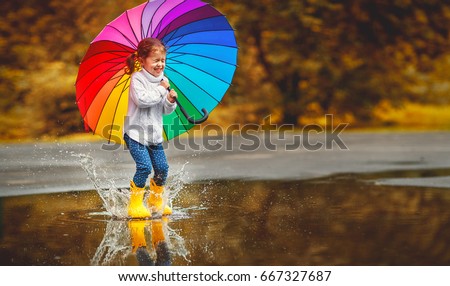 Happy funny ba child by girl with a multicolored umbrella jumping on puddles in rubber boots and laughing