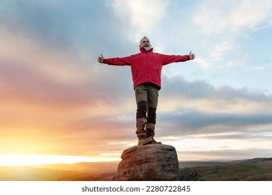 happy funny bearded  Man in red  jacket relaxing alone on the top of  mountain  at sunrise. Travel  Lifestyle concept The national park Peak District in England
