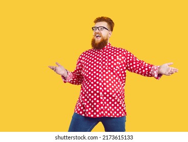 Happy funny cheerful fat young man wearing red white polka dot shirt and glasses isolated on solid yellow colour background looks away, sees something unusual and opens his mouth in surprise