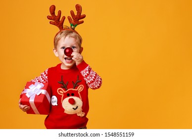 Happy Funny Child Boy In Red Christmas Reindeer Costume With Gift On Yellow Colored Background
