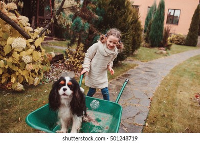 Happy Funny Child Girl Riding Her Dog In Wheelbarrow In Autumn Garden, Candid Outdoor Capture, Kids Playing With Pets