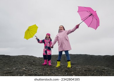 Happy funny children with pink and yellow umbrella outdoors. Girls wearing pink raincoat and enjoying rainfall. Kid playing on the nature outdoors