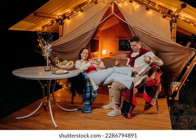 Happy funny couple relaxing in glamping on summer evening and drinking wine near cozy bonfire. Luxury camping tent for outdoor recreation and recreation. Lifestyle concept