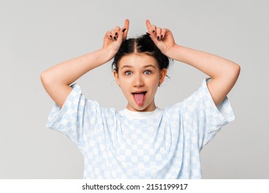 Happy funny dark-haired teen girl shows bull horns on head and stuck out her tongue, fooling around and playing, stands in trendy t shirt over light grey background