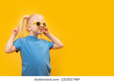 Happy funny fair-haired cheerful little girl wear sunglasses and blue t-shirt on yellow background. copy space