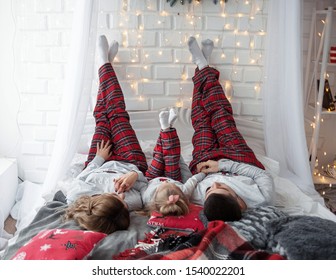 Happy Funny Family With Their Daughter In The Bedroom In The Same Beautiful Christmas Pajamas With Their Raised Legs Opposite Wall While Relaxing At Home. Christmas Morning Celebration.