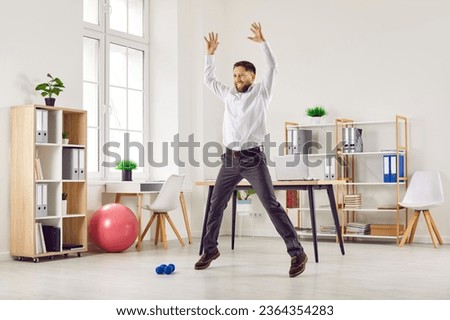 Happy funny fit smiling young business man or corporate employee doing star jumps during a quick fitness workout at work in the office. Sport, physical activity, healthy lifestyle concept