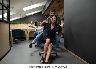 Happy Funny Friendly Diverse Coworkers Having Fun Racing Riding On Chairs In Modern Office, Asian Employee Laughing Enjoying Team Building Activity At Work Break Laughing Playing At Workplace