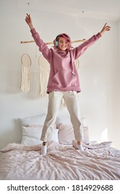 Happy funny funky hipster gen z woman with pink hair wears hoodie headphones listening pop music playlist or podcast, jumping, dancing, having fun standing on bed in cozy bedroom.