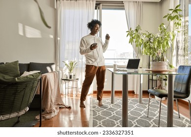 Happy funny gen z hipster African American teen guy wearing headphones dancing at home, listening music on mobile phone, having fun feeling funky moving in living room, authentic shot. - Powered by Shutterstock