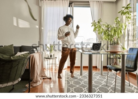 Happy funny gen z hipster African American teen guy wearing headphones dancing at home, listening music on mobile phone, having fun feeling funky moving in living room, authentic shot.