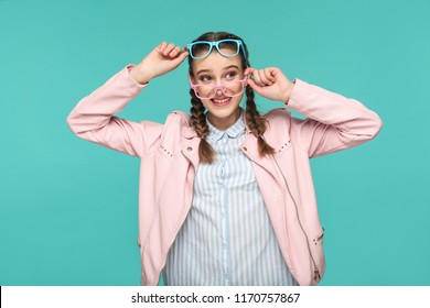 Happy funny girl in casual or hipster style, pigtail hairstyle, standing, holding two blue and pink glasses and looking away with toothy smile, Indoor studio shot, isolated on blue or green background