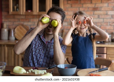 Happy Funny Girl And Dad Cooking Salad In Kitchen, Playing And Having Fun, Holding Fresh Vegetables At Eyes Making Glasses, Goggles, Enjoying Home Activities, Keeping Healthy Organic Food Diet