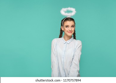 Happy funny girl in striped blue shirt and pigtail hairstyle, standing with halo on her head and looking away with smile and dreaming face, Indoor studio shot, isolated on blue or green background