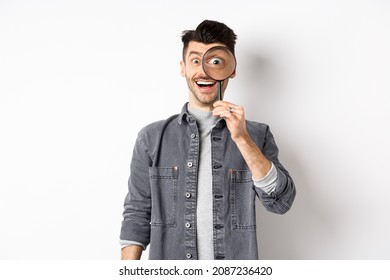 Happy funny guy look through magnifying glass with big eye, checking out interesting deal, searching something, standing on white background