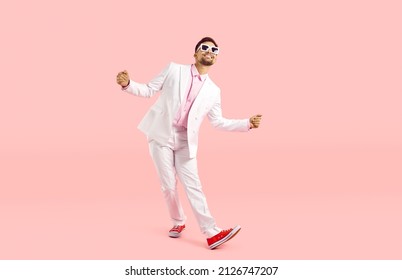 Feliz tipo gracioso con traje blanco bailando aislado con fondo rosa pastel. Captura completa de un alegre hombre despreocupado con traje moderno, zapatillas de deporte y gafas bailando y divirtiéndose en el estudio