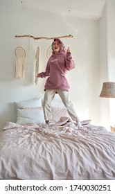 Happy funny hipster teenage girl with pink hair wear hoodie and headphones listening radio pop music playlist podcast, singing song jumping dancing standing on bed in cozy hygge bedroom. Vertical