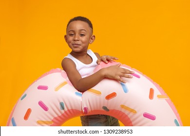 Happy Funny Little Boy Of African Appearance Having Fun, Posing At Yellow Wall With Inflatable Pink Doughnut Circle, Going To Beach. Adorable Black Male Child Carrying Swimming Ring, Enjoying Summer