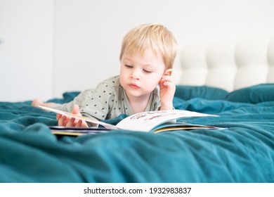 Happy funny little boy in pajamas reading book lying in his parents bed.