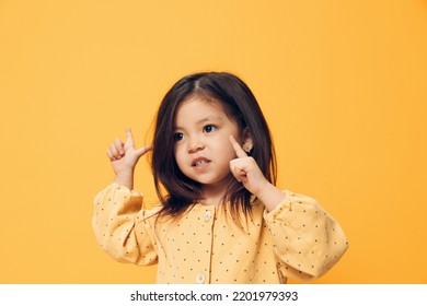 A Happy, Funny Little Girl Of Preschool Age Stands On An Orange Background In A Trendy Dress And Points Her Fingers At The Empty Space Of The Background.