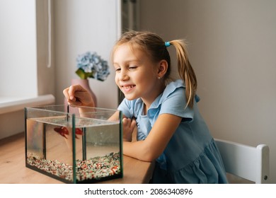 Happy funny little girl watching little goldfish in aquarium, tapping on glass, attracting attention. Portrait of pretty cheerful preschool kid playing with gold fish at home.
