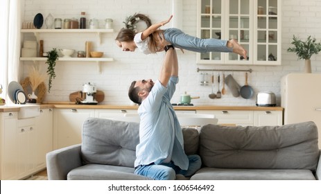 Happy funny little kid girl flying in fathers arms family having fun in modern studio apartments, loving single daddy holding lifting cheerful pre-school daughter playing plane enjoy playtime at home