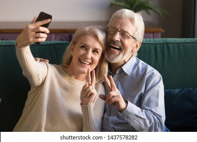 Happy funny mature senior couple taking selfie looking at smartphone, cheerful elder old grandparents having fun holding phone make snapshot self portrait laughing showing peace sign sitting on sofa