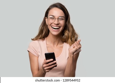 Happy funny millennial woman euphoric celebrating mobile win victory triumph holding phone, cheerful excited girl laughing having fun using cellphone apps isolated on white studio blank background