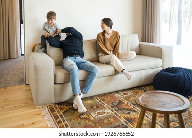 Happy Funny Parents And Cute Little Child Son Laughing In Modern Living Room. Joyful Parent Dad Sitting At The Sofa And Catching His Son While Playing With Boy Together. Fun And Home Concept