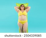 Happy funny plus size woman in sunglasses and in summer yellow bikini lying on inflatable mattress isolated on a studio blue background. Fat girl going to swim on a beach. Vacation concept.