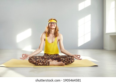 Happy Funny Relaxed Smiling Skinny Man With Hippie Style Long Hair In Nerd Glasses, Yellow Retro Headband, Tank Top And Leopard Print Leggings Meditating Eyes Closed In Lotus Yoga Pose On Exercise Mat