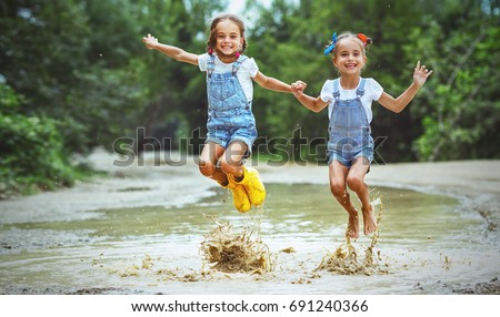 Happy funny sisters twins  child by girl jumping on puddles in rubber boots and laughing
