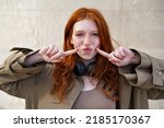 Happy funny teen stylish cool redhead girl standing on urban wall background looking at camera. Portrait of pretty smiling teenage girl with red hair and freckles having fun laughing outdoor.