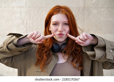 Happy funny teen stylish cool redhead girl standing on urban wall background looking at camera. Portrait of pretty smiling teenage girl with red hair and freckles having fun laughing outdoor.