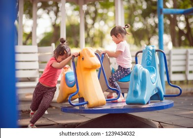 Happy Funny Two Little Children Playing With Merry Go Around At The Playground. Image Of Emotional Cheerful Adorable Asian Kids Having Fun In Summer Of Outdoors. Education Activity For Kid Concept.