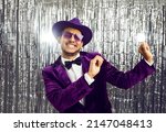 Happy funny young black man wearing tacky unfashionable purple velvet suit, hat and sunglasses dancing at party. Goofy ethnic guy dancing on concert show stage with shiny foil fringe studio background