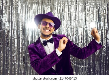 Happy funny young black man wearing tacky unfashionable purple velvet suit, hat and sunglasses dancing at party. Goofy ethnic guy dancing on concert show stage with shiny foil fringe studio background - Shutterstock ID 2147048413